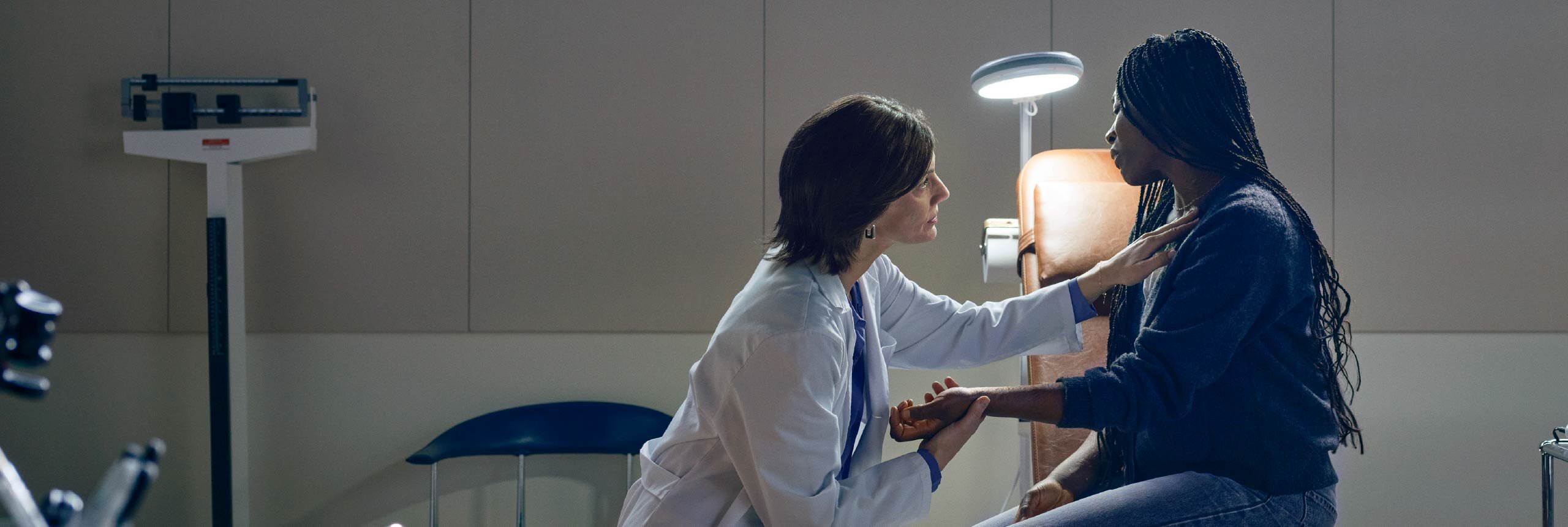 Doctor speaking with a seated patient in a medical exam room, with a hand on the patient’s shoulder and holding the patient’s hand.